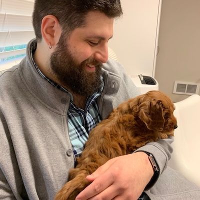 Alignment Therapy owner and OCD / PTSD / anxiety therapist Daniel Silvermint looking down at a red puppy he's holding with his left arm. Daniel is a bearded middle-aged white man wearing a light blue checkered shirt and a light grey fleece.